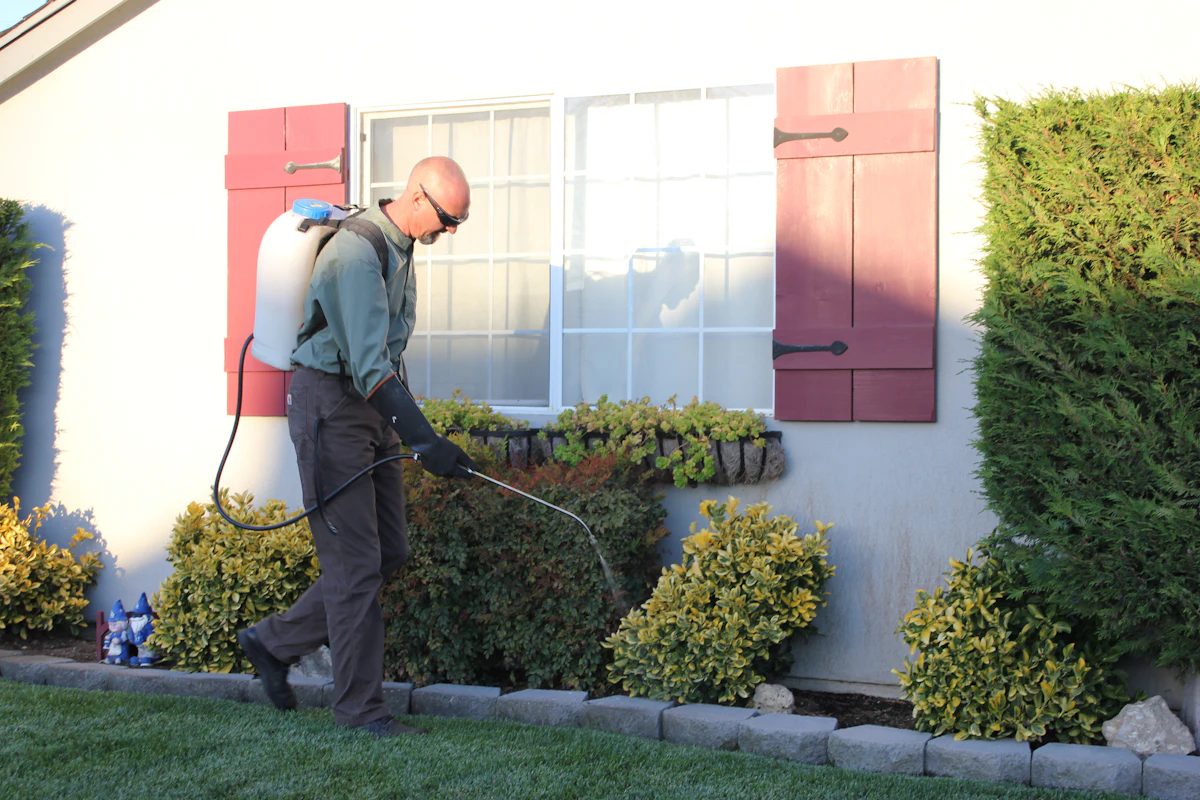 A pest control technician in a uniform and gloves, carrying a backpack sprayer, treats the planting beds along the front wall of a house.